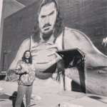 Steven Adams posing next to his murial on a brick wall